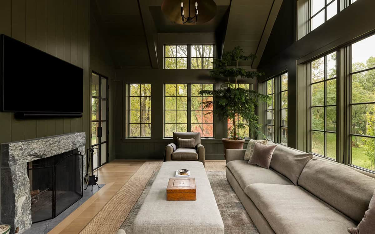 Family room with deep green vertical paneling, a vaulted ceiling, a chunky stone fireplace, a gray sofa, a linen ottoman and walls of windows