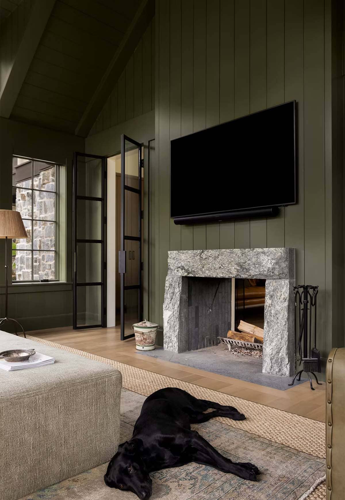 Rough-cut stone fireplace surround against green paneling, with steel-framed glass doors and a black Labrador lying on the rug