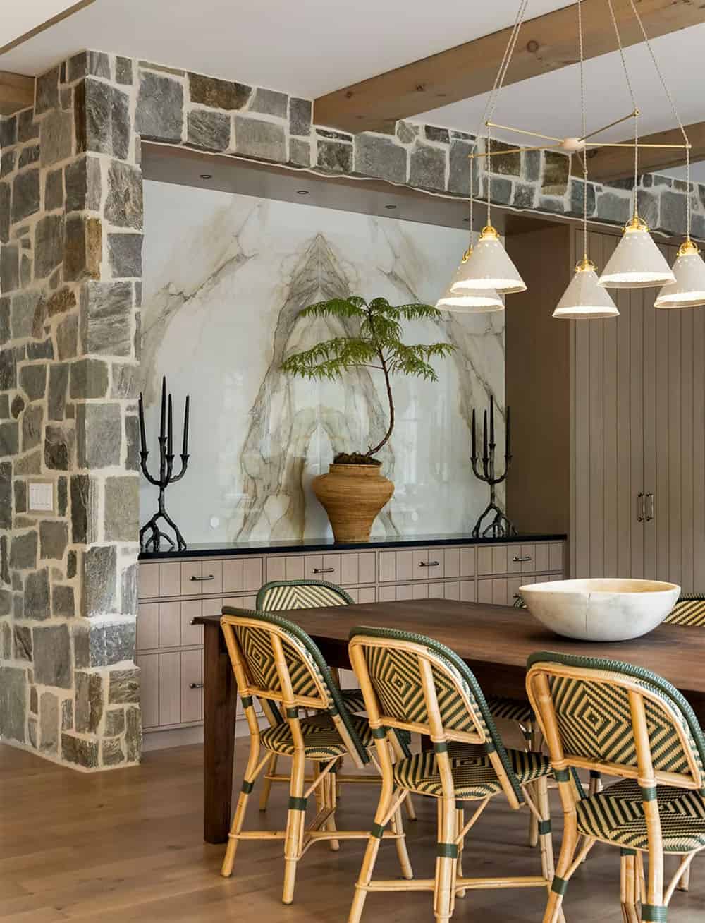 Marble-backed buffet niche with black branch candelabras and a delicate tree in a woven vessel, with cone pendants over rattan dining chairs