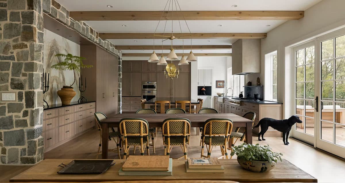 Dining area flowing into an oak kitchen under wood ceiling beams, with a black Labrador standing by French doors to the deck
