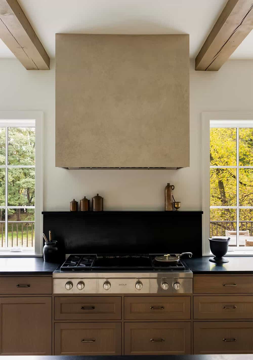 Range wall with a textured plaster hood, a Wolf rangetop, a black stone backsplash with a display ledge and windows on either side