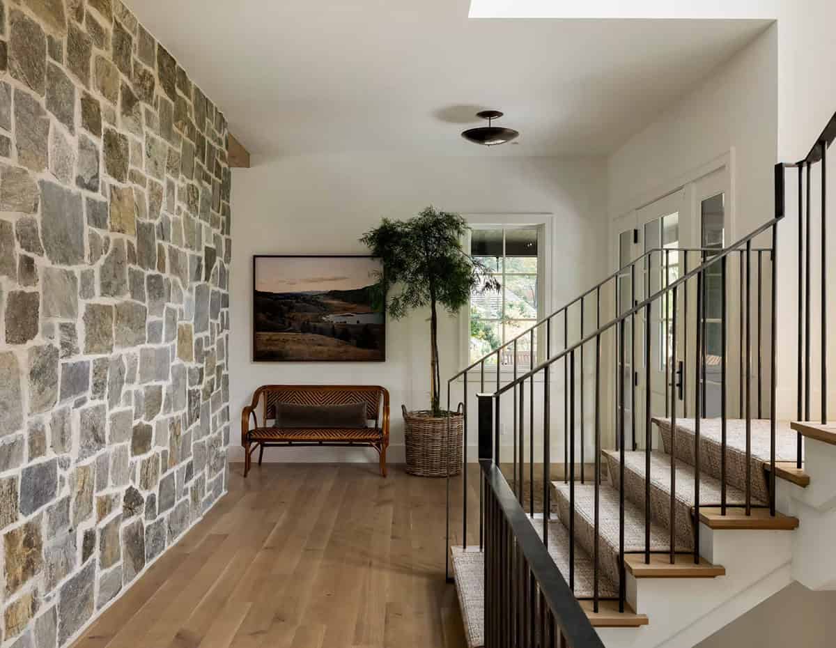 Entry hall with a full-height stone wall, a vintage rattan bench, a landscape painting, a potted tree and a carpeted staircase with a black metal railing