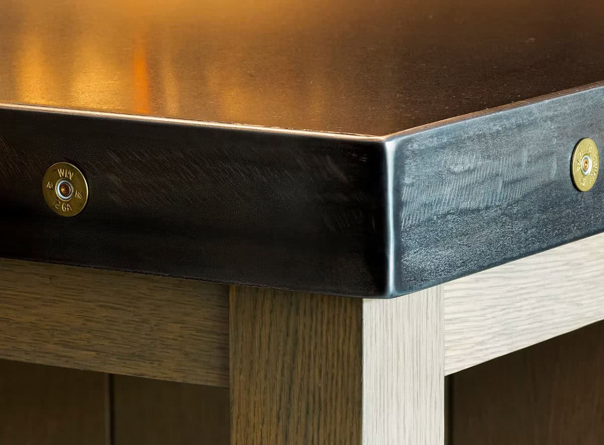 Close-up of a blackened steel bar countertop edge with brass shotgun shell heads set in as rivets above a white oak base