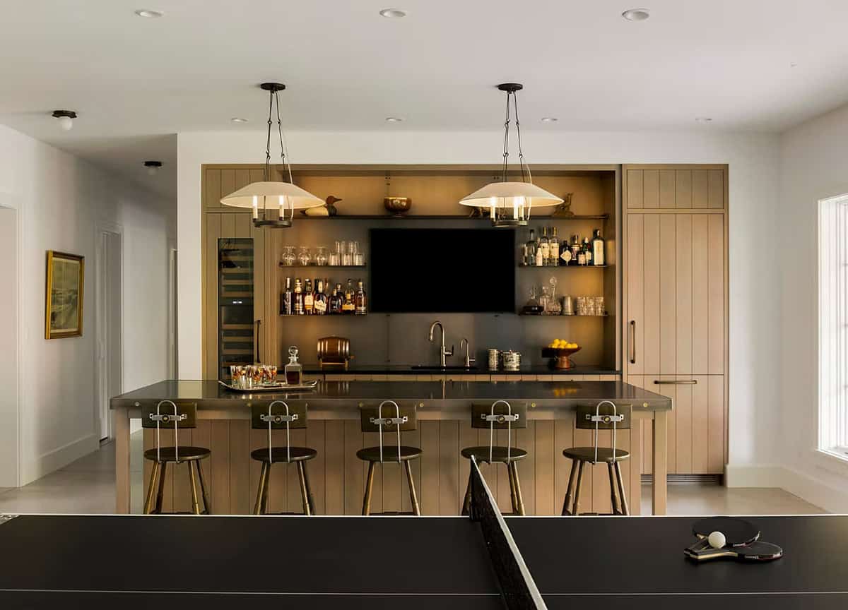 Lower-level bar with light oak paneling, open shelves of bottles and glassware, metal barstools and a ping-pong table in the foreground