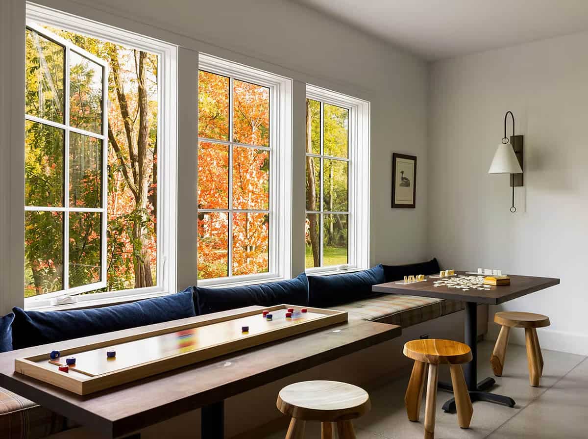 Game nook with a built-in banquette of plaid cushions and navy pillows, a tabletop shuffleboard, dominoes and wood stools by windows of fall color