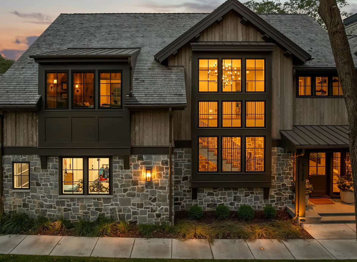 Front facade at dusk with lit windows revealing a branch chandelier and staircase behind a two-story window bay above a stone base