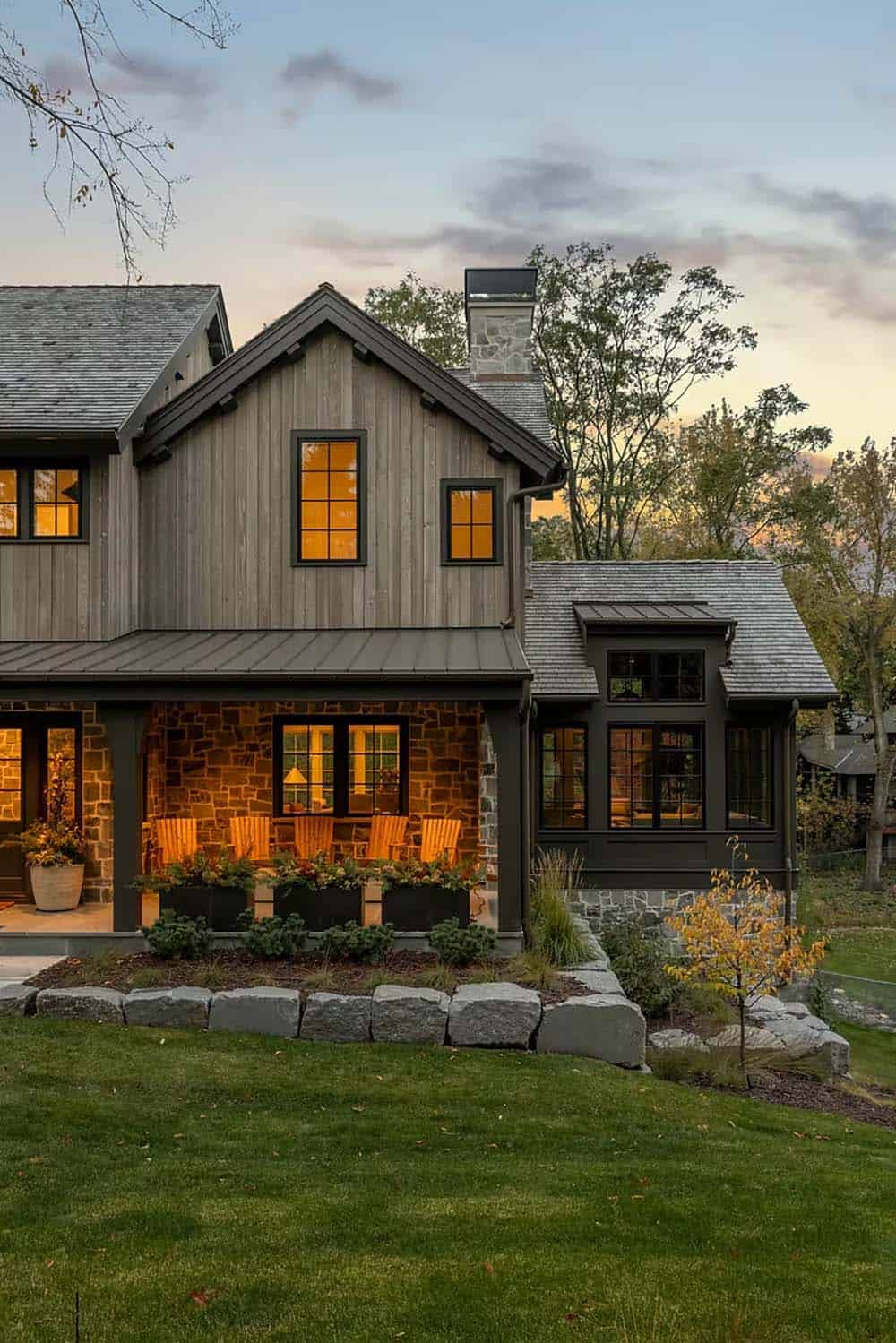 Front porch and stone chimney at twilight, with glowing windows, vertical wood siding and granite boulders edging the lawn