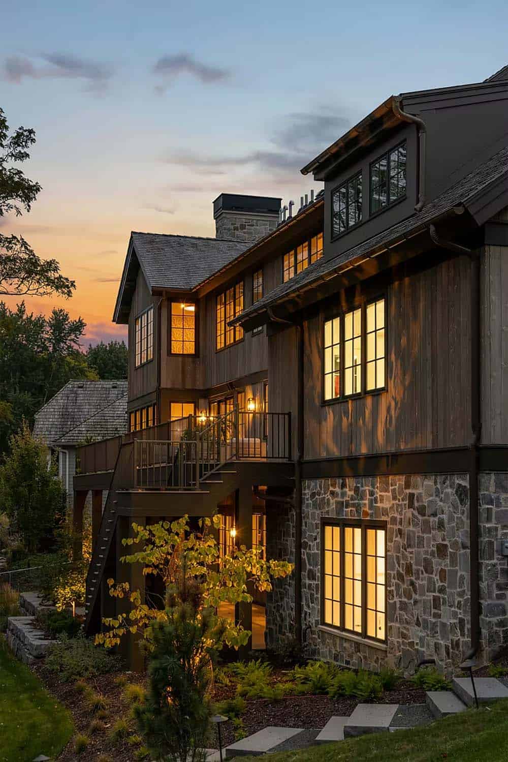 Rear of the home at sunset with a lit multi-level deck, weathered wood siding, a stone base and landscape lighting along stone steps