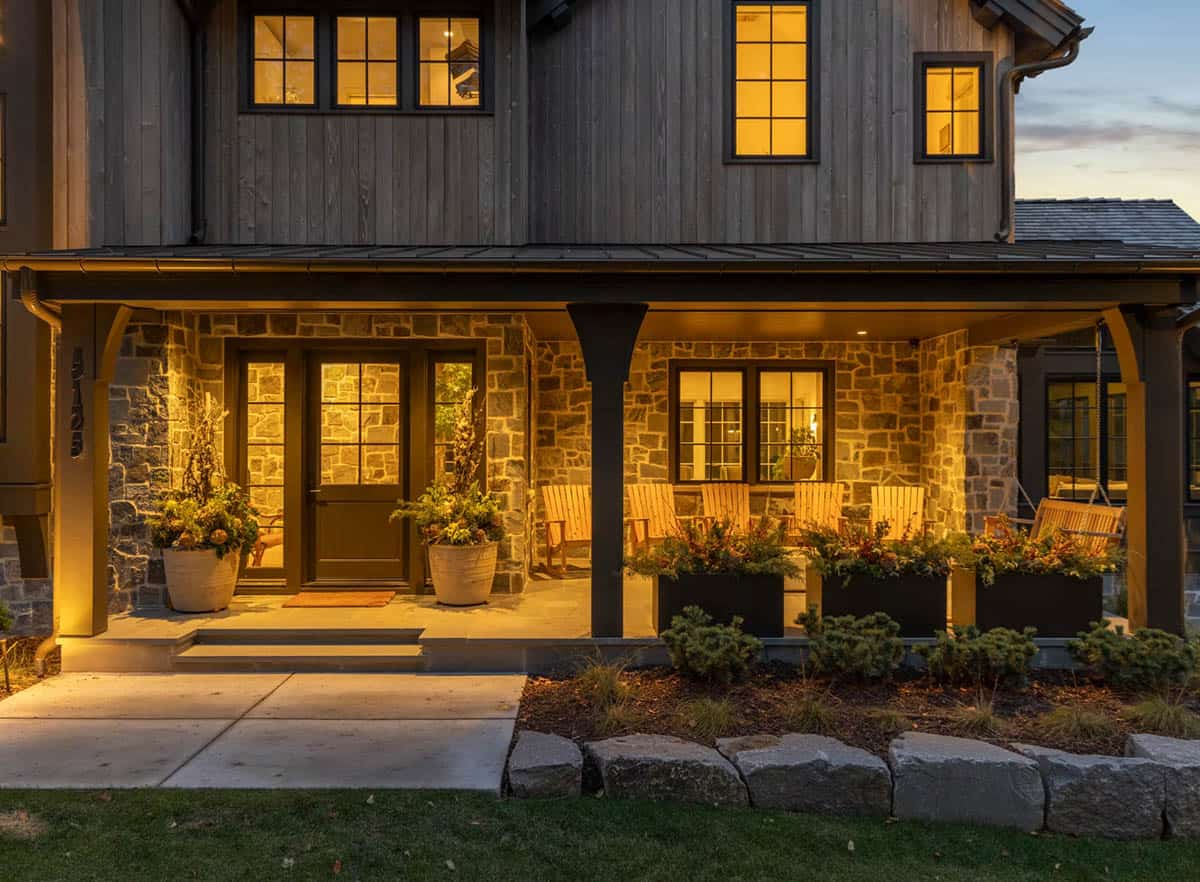 Front porch at dusk glowing with warm light, with Adirondack chairs, a porch swing, seasonal planters and a stone wall
