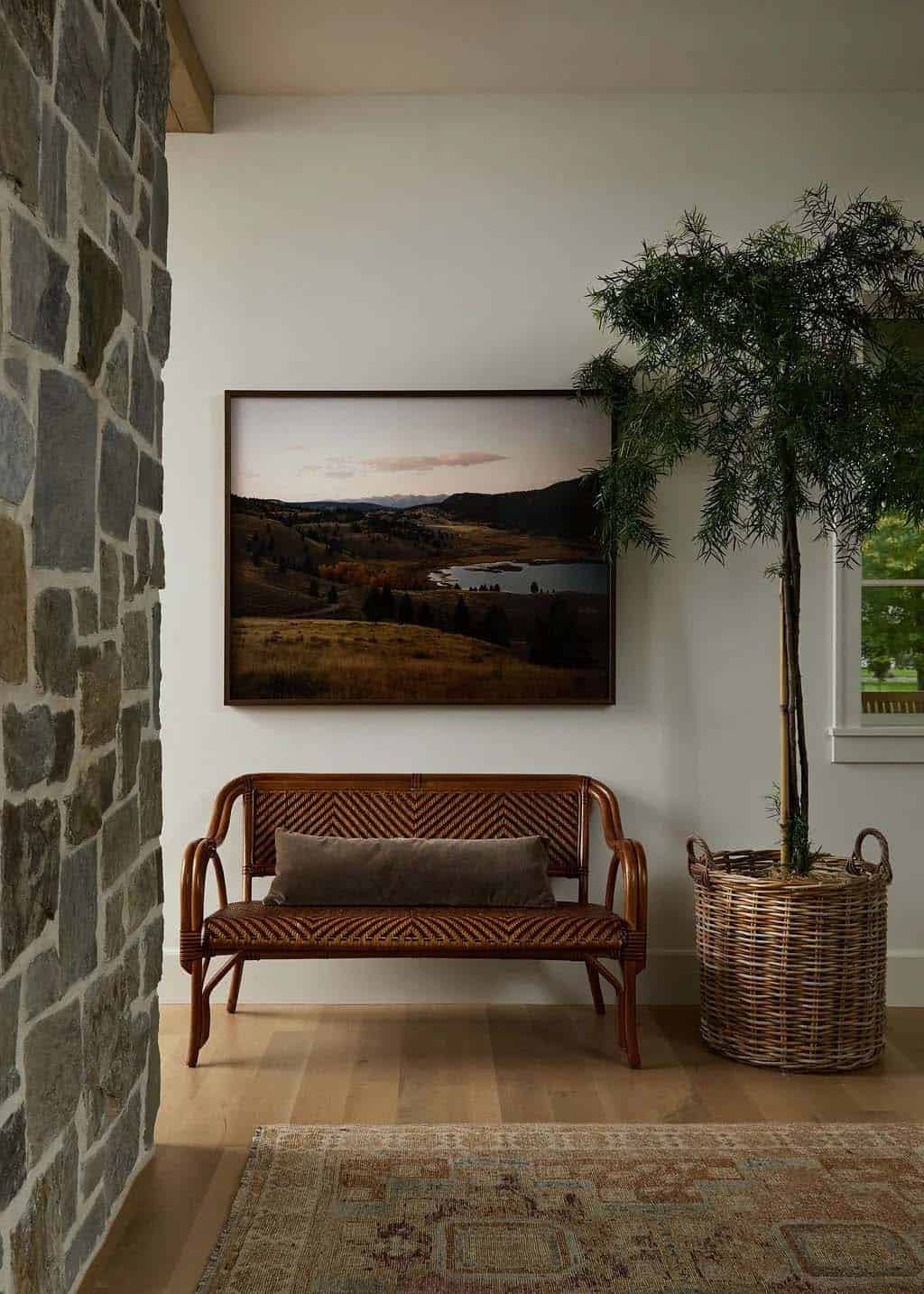 Entry nook with a woven rattan bench, a framed mountain landscape photograph and a potted tree in a wicker basket beside a stone wall