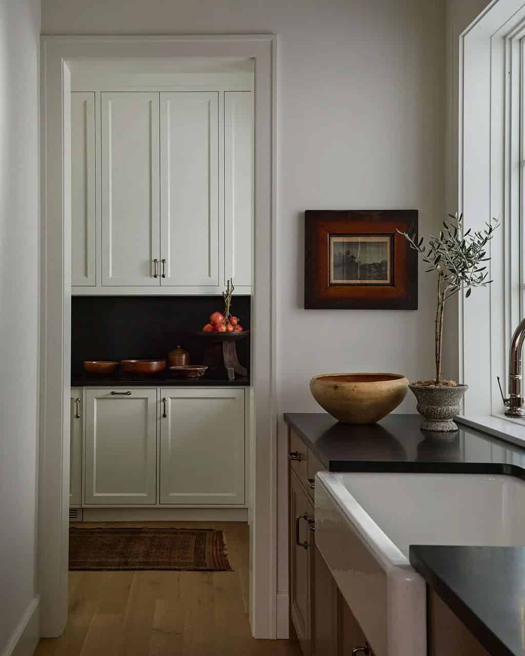 View from the kitchen's farmhouse sink into a walk-in pantry with cream cabinets, a black backsplash and wooden bowls