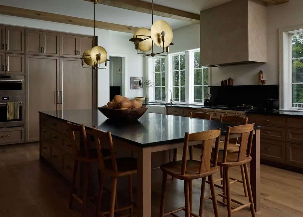 Kitchen island with a black countertop, carved wood counter stools and brass disc pendants, with oak cabinetry and a plaster range hood beyond