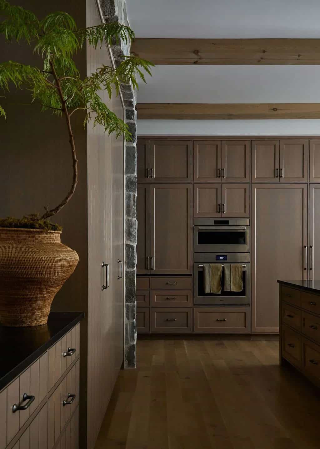 Floor-to-ceiling oak cabinetry with paneled refrigerator doors and double wall ovens beside a stone pier and a tree in a woven pot