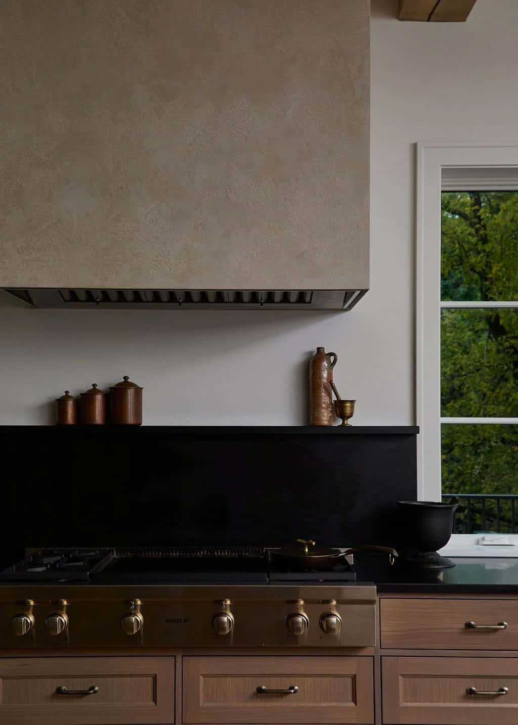 Plaster range hood above a black stone backsplash ledge with copper canisters, a Wolf rangetop and a window to the trees