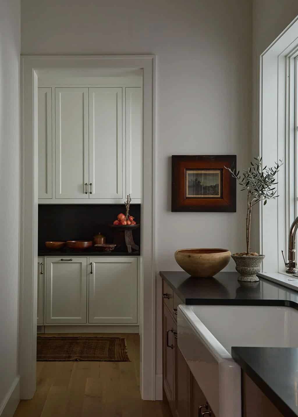 Walk-in pantry with cream cabinets and a black stone backsplash seen through a doorway beside a framed landscape and an olive tree