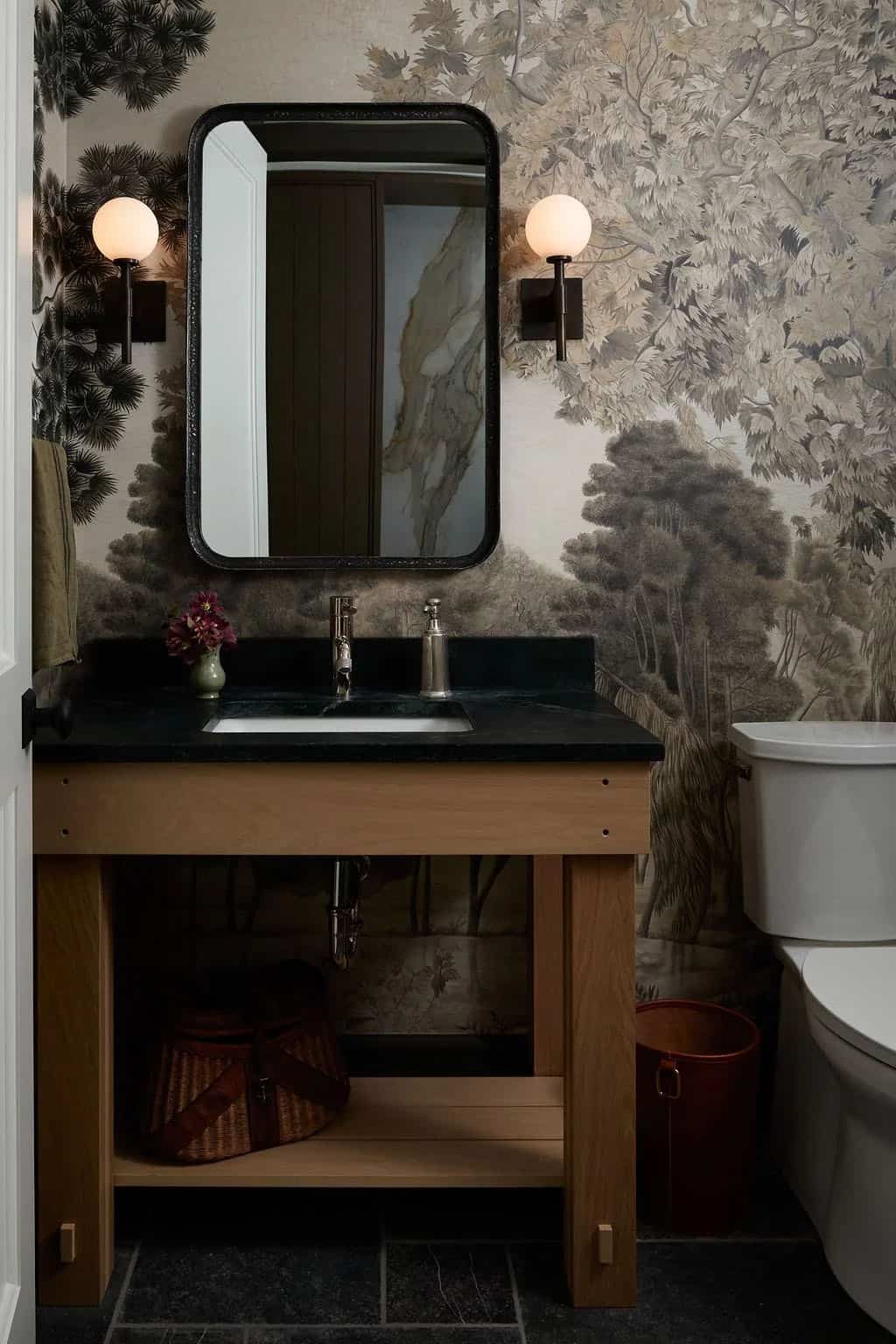 Powder room with a scenic tree mural, a black-framed mirror, globe sconces and an open oak vanity with a black stone top and a vintage fishing creel