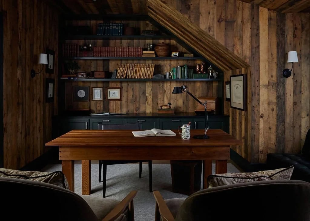 Barnwood-paneled home office with a wood desk, a black task lamp and dark green built-in shelves filled with leather-bound books