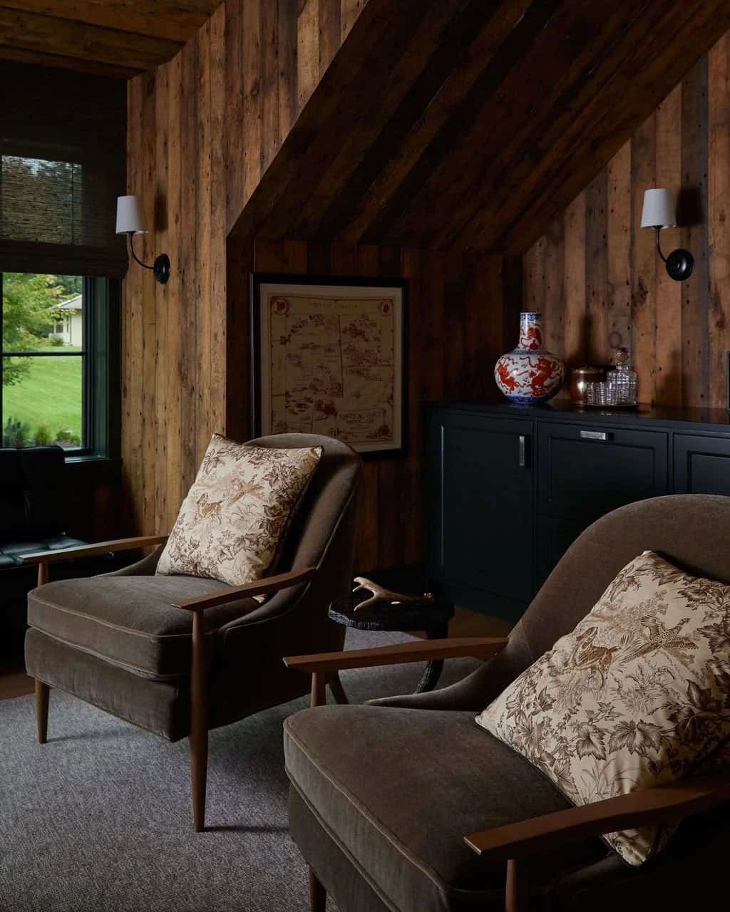 Two mohair lounge chairs with toile pillows in a barnwood office, beside dark green cabinets holding a blue-and-white vase