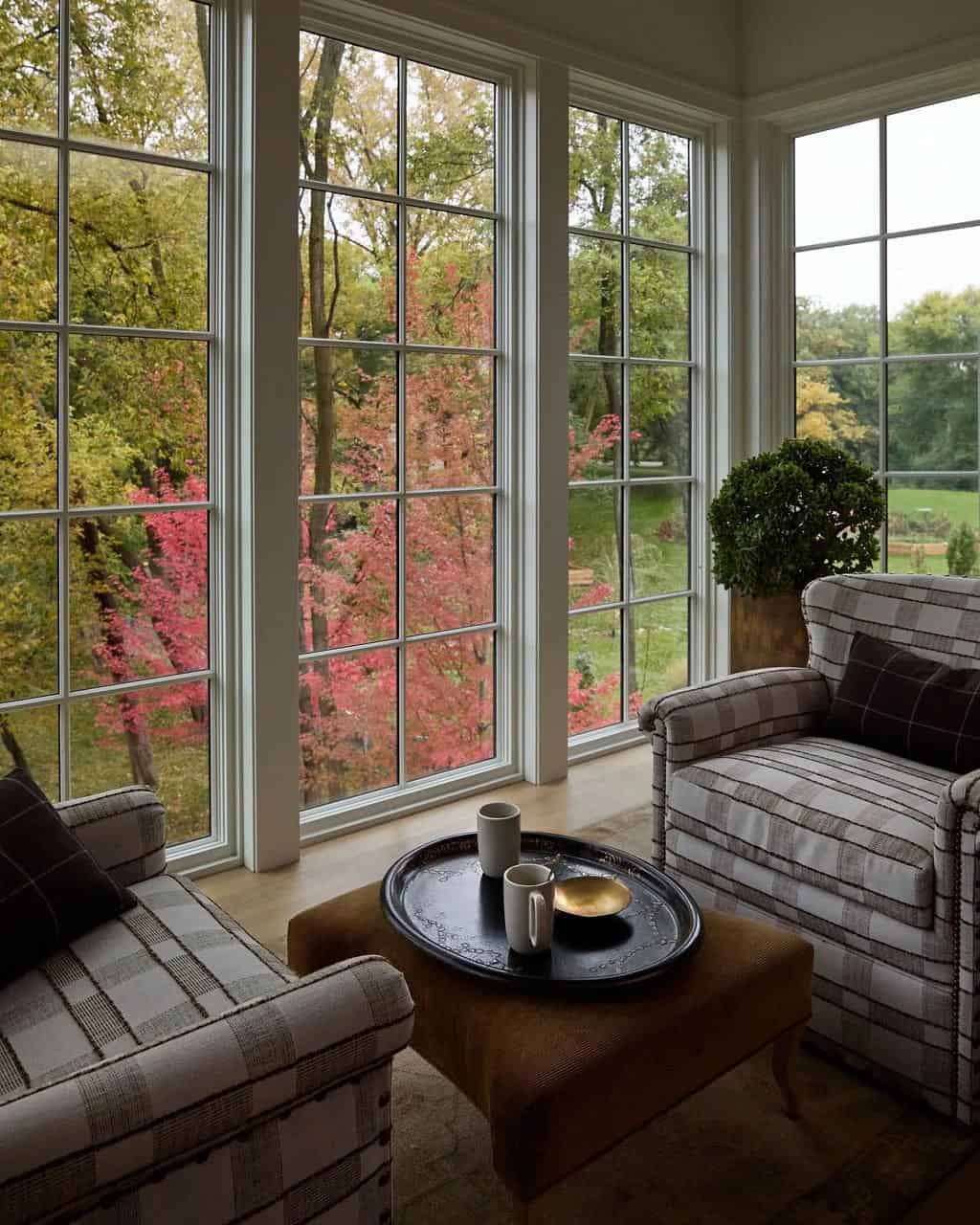 Two plaid armchairs and a corduroy ottoman with a black tray beside floor-to-ceiling windows looking out to red and yellow fall trees