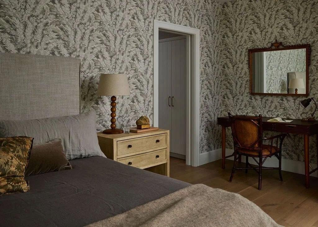 Bedroom with leafy botanical wallpaper, a linen upholstered bed, an oak nightstand with a beaded wood lamp and a writing desk with a cane chair
