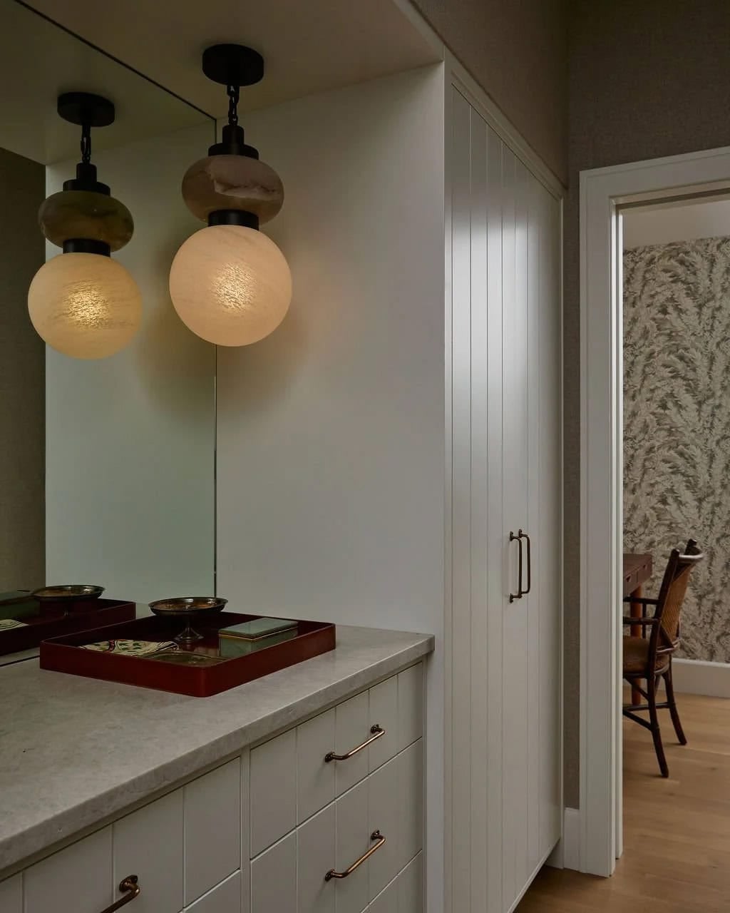 Dressing area with alabaster pendant lights, a gray stone countertop, white paneled cabinetry and a doorway to a wallpapered bedroom
