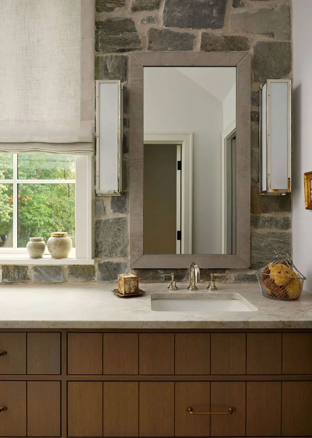 Primary bathroom sink with a wood-framed mirror and nickel sconces on a natural stone wall beside a window with a linen Roman shade