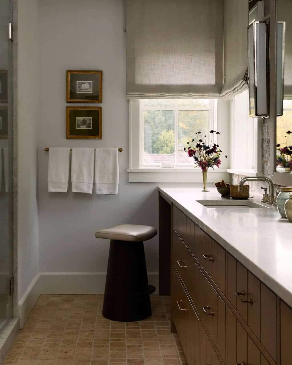 Primary bathroom with a long oak vanity and stone countertop, a sculptural stool under a linen Roman shade and towels on a brass bar