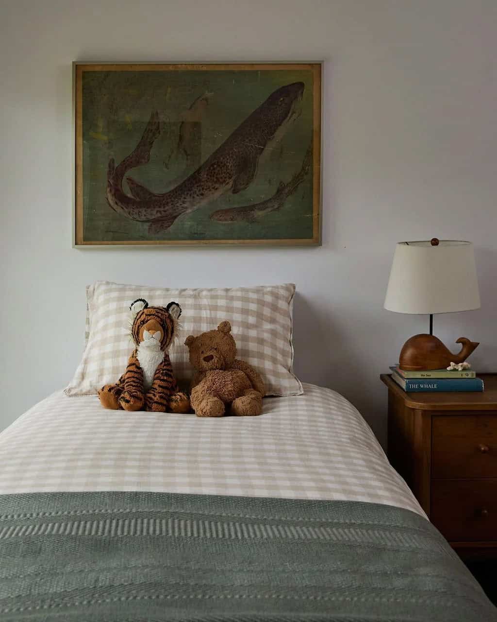 Twin bed with gingham bedding, a stuffed tiger and teddy bear, a vintage shark print and a carved wooden whale lamp on a nightstand