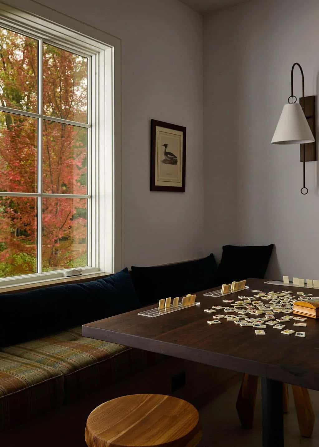 Game nook with a navy banquette and plaid cushions, a dark wood table set with dominoes and a window framing red fall leaves