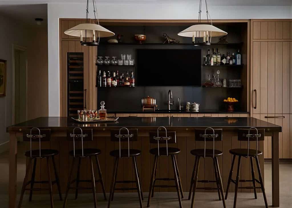 Lower-level bar with an oak-paneled island, six metal-back barstools, open shelves of bottles and glassware, and a wine fridge