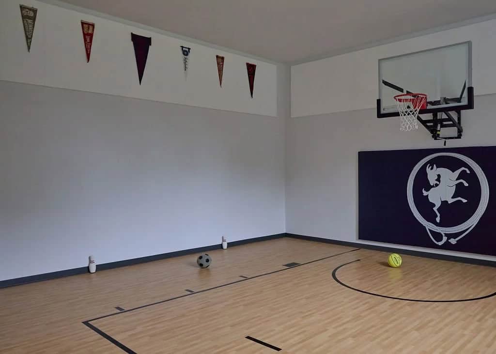 Indoor sport court with a basketball hoop, wood-look flooring, college pennants on the walls and a navy wall pad with an animal logo