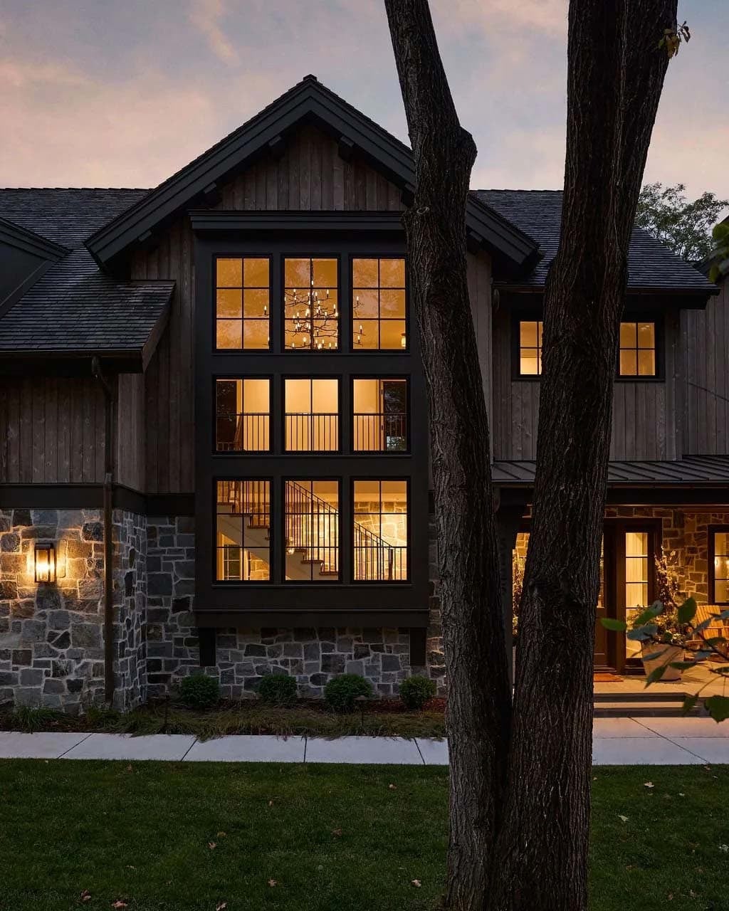 Two-story window bay glowing at dusk, revealing a branch chandelier and staircase behind a tall tree trunk and a stone facade