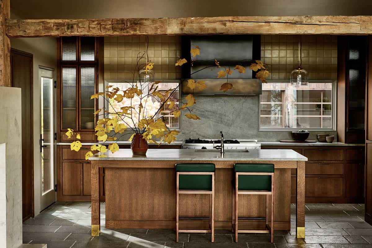 Oak kitchen with brass-capped island, green leather stools, bronze range hood and glazed tile backsplash