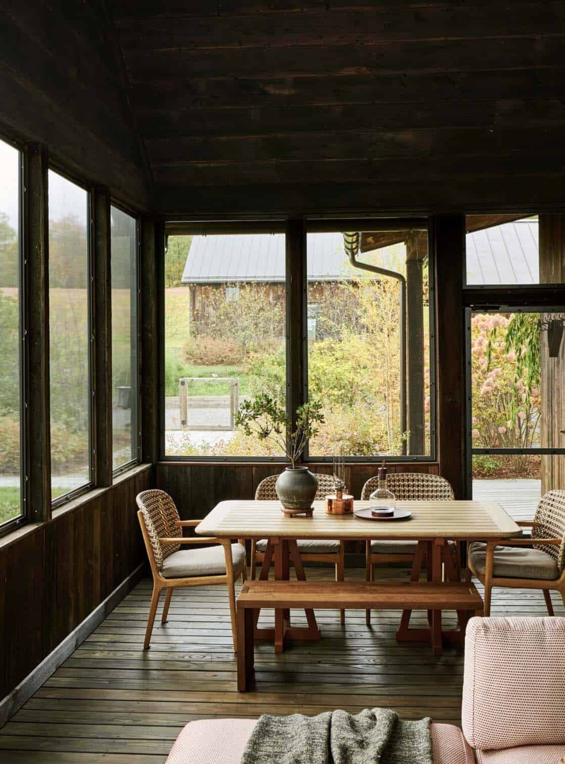Screened porch with dark wood walls, a teak dining table, woven chairs and garden views