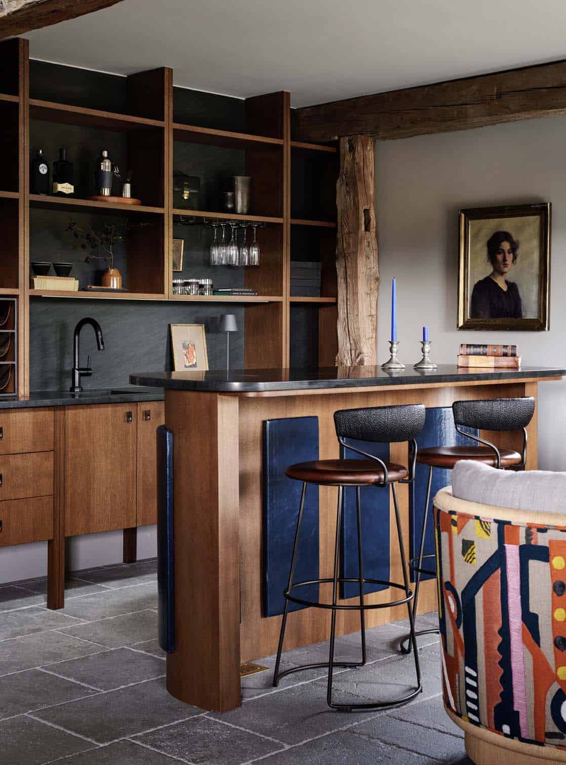 Bar with oak open shelving, slate backsplash, curved island with blue leather panels and black stools