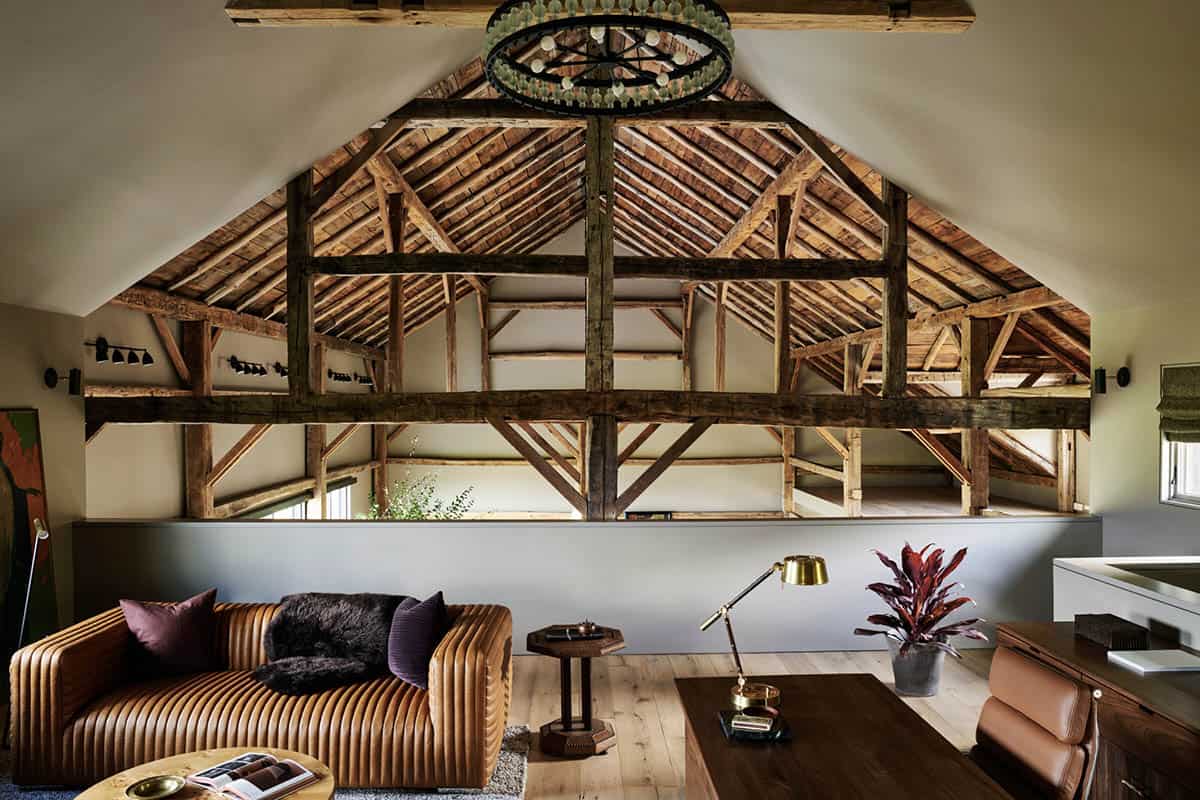 Loft office overlooking the barn's open timber frame, with cognac leather sofa, brass lamp and wood desk