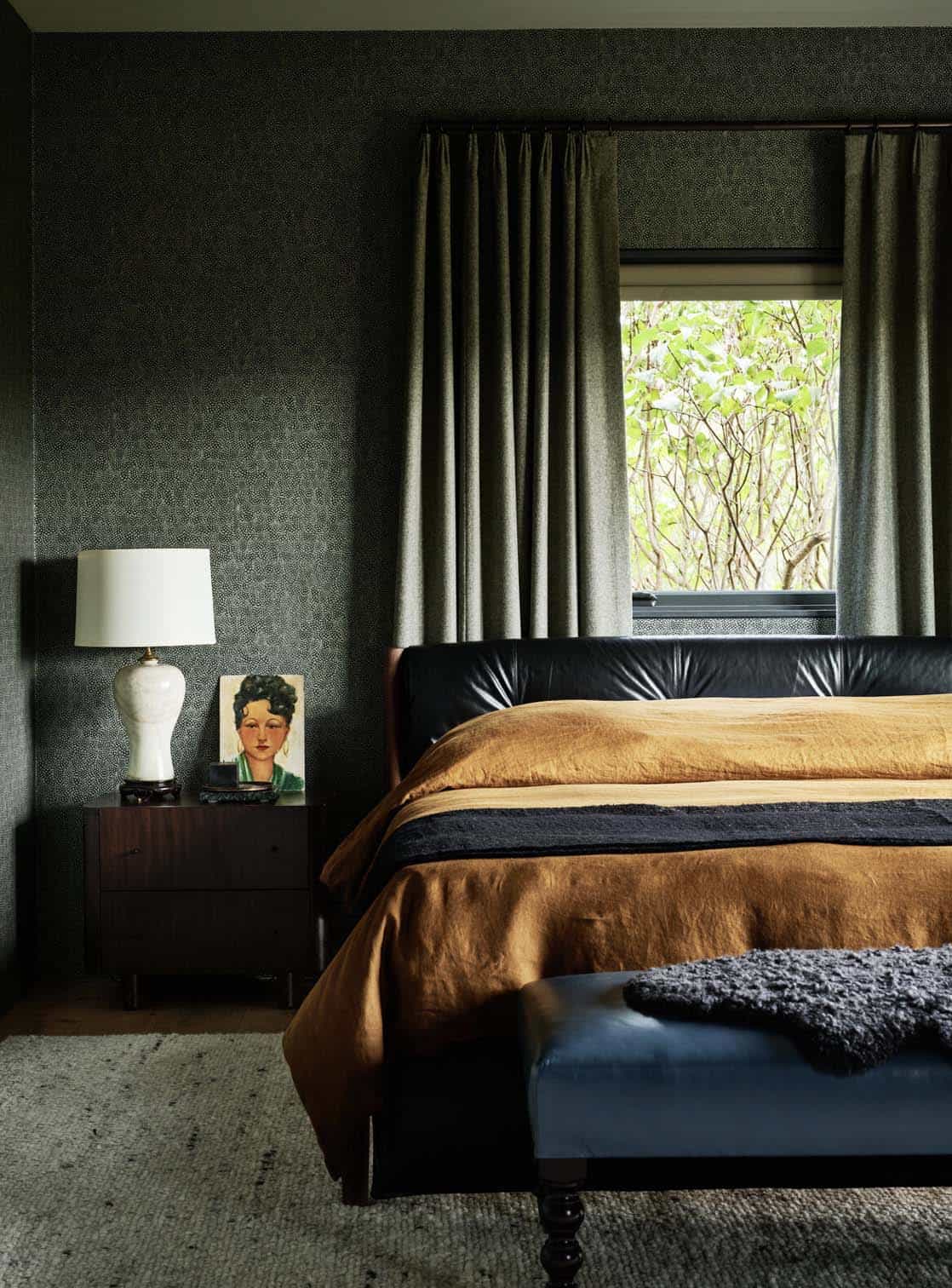 Bedroom with dark patterned wallpaper, black leather bed, ochre linen bedding and a blue leather bench