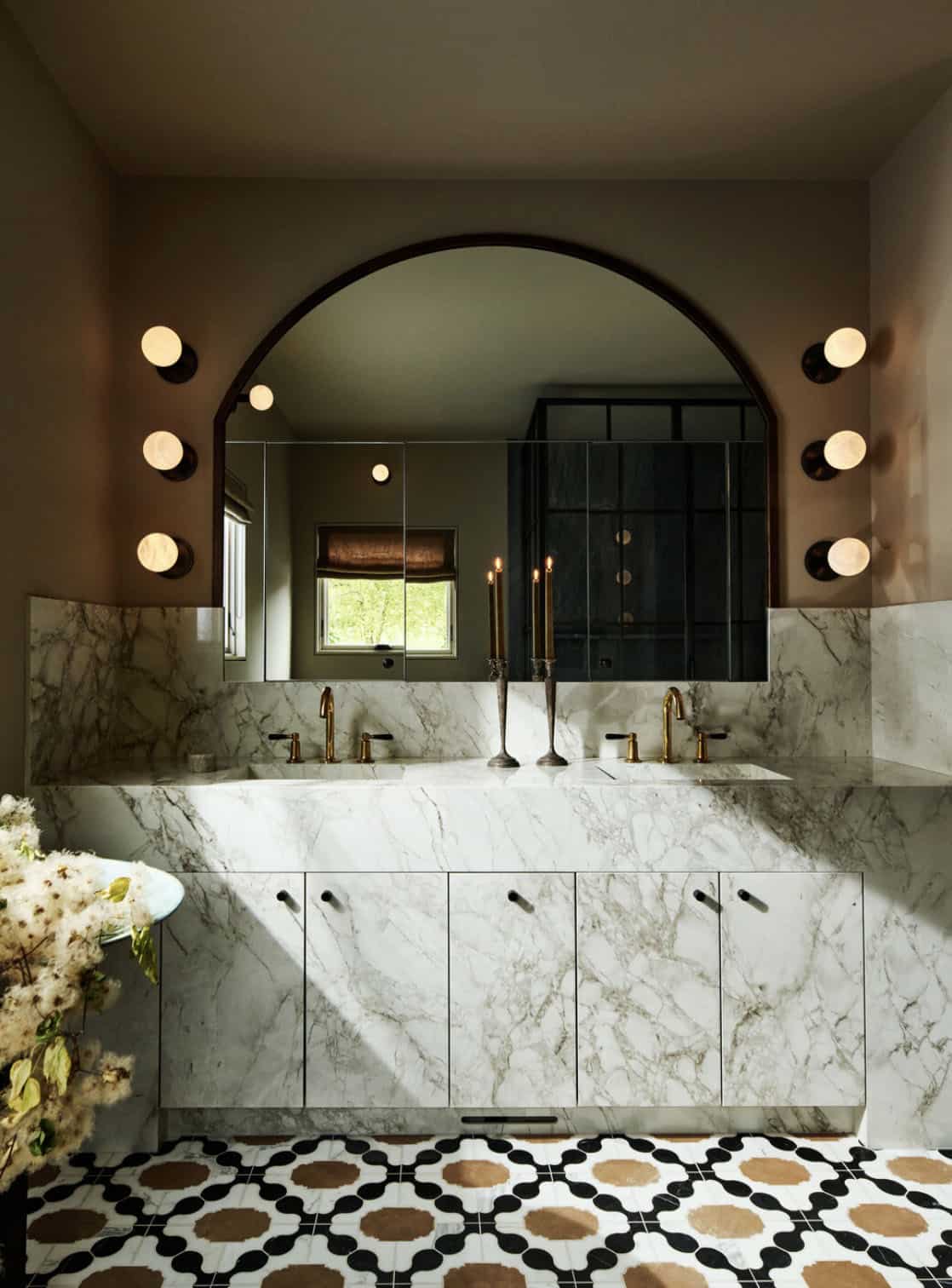 Bathroom with porcelain slab double vanity, arched mirror, globe sconces and black, white and tan mosaic floor