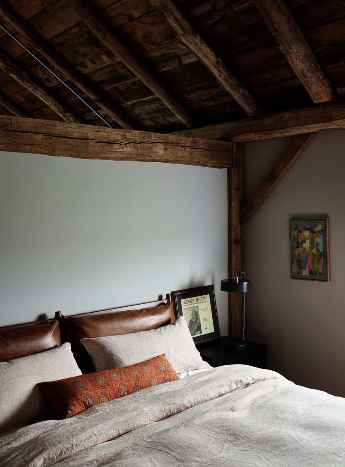 Bedroom under dark timber ceiling beams with a leather headboard, linen bedding and a rust patterned pillow