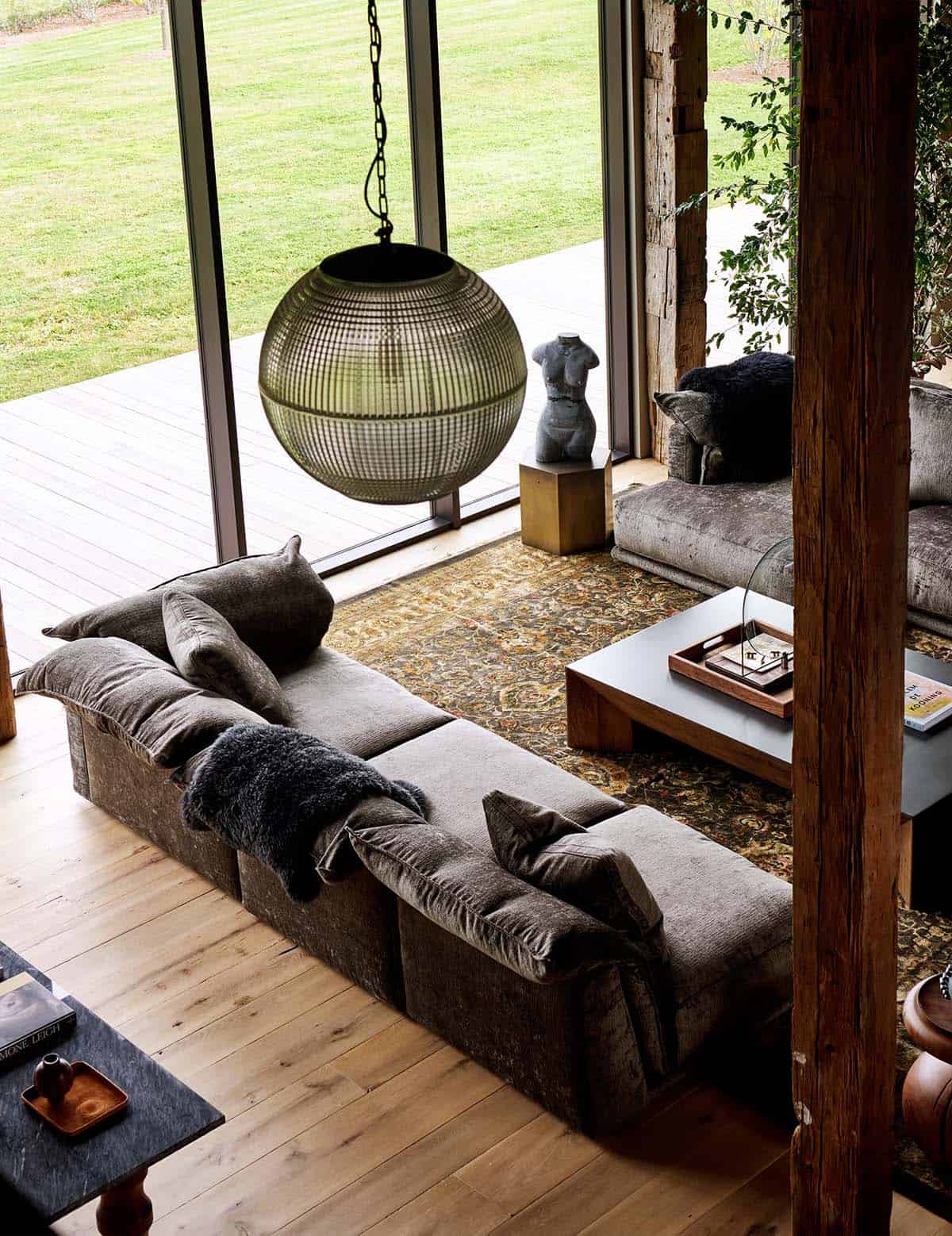 Overhead view of gray velvet sofa, glass globe pendant and torso sculpture beside windows to the lawn