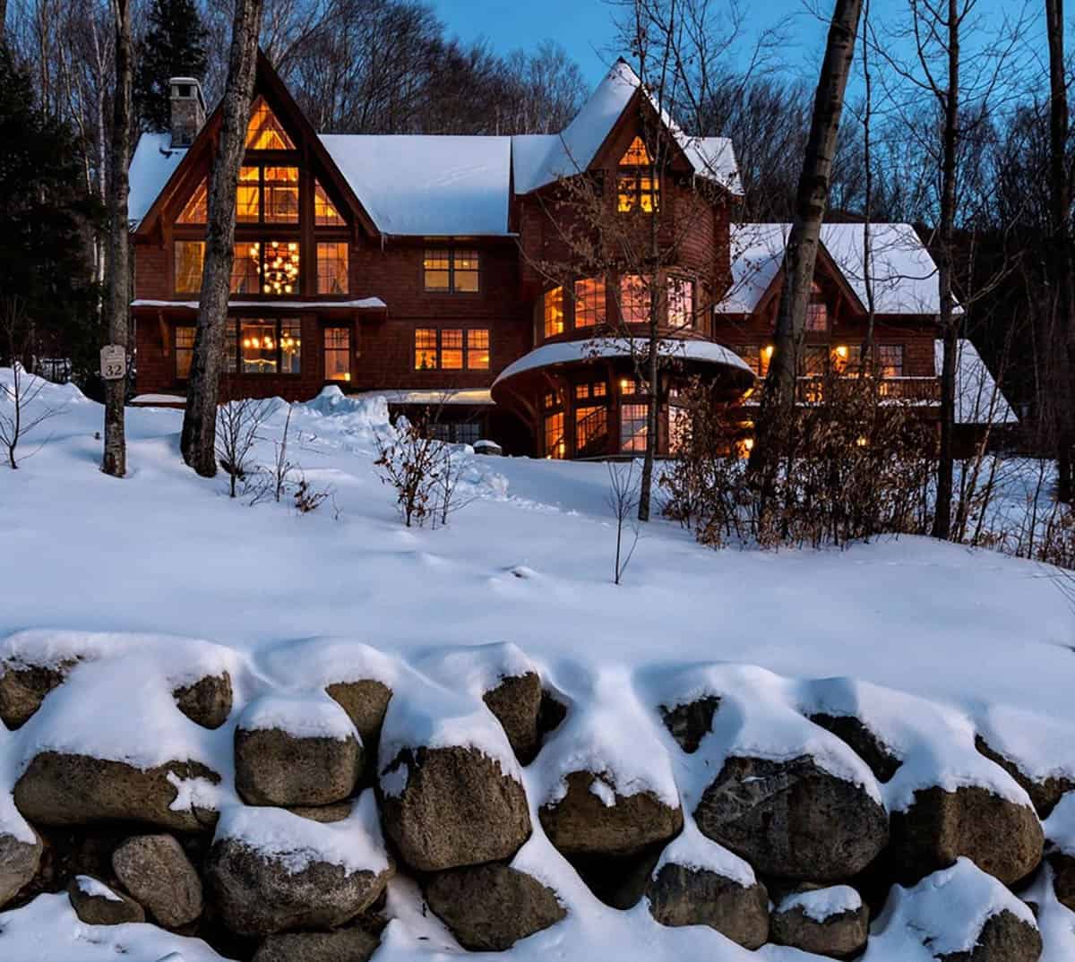 Shingle mountain home glowing at twilight behind a snow-capped boulder wall