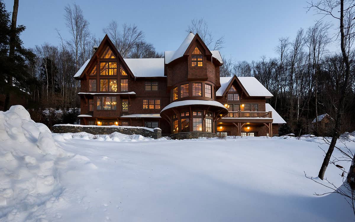 Shingle-clad mountain home at dusk with steep snow-covered gables, a curved turret and glowing windows above a snowy lawn