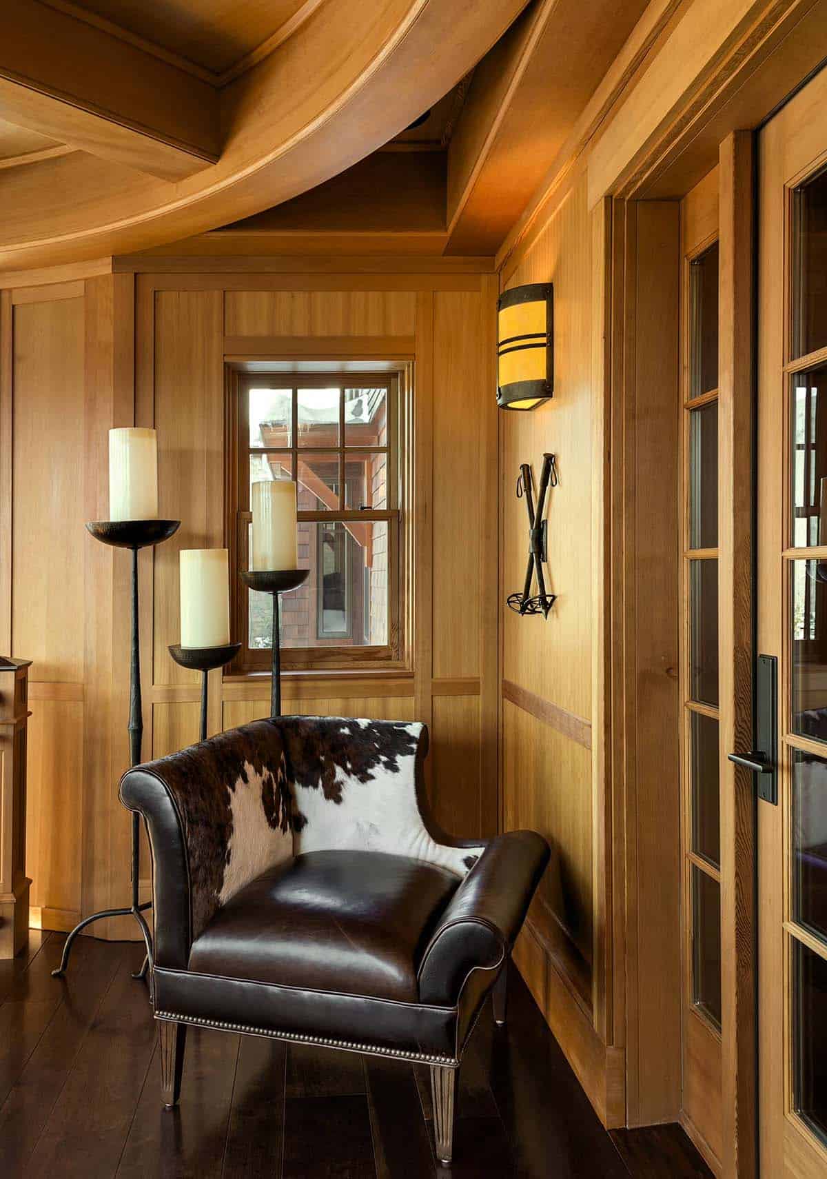 Cowhide and leather chair beside iron candle stands, with vintage ski poles mounted on a wood-paneled wall