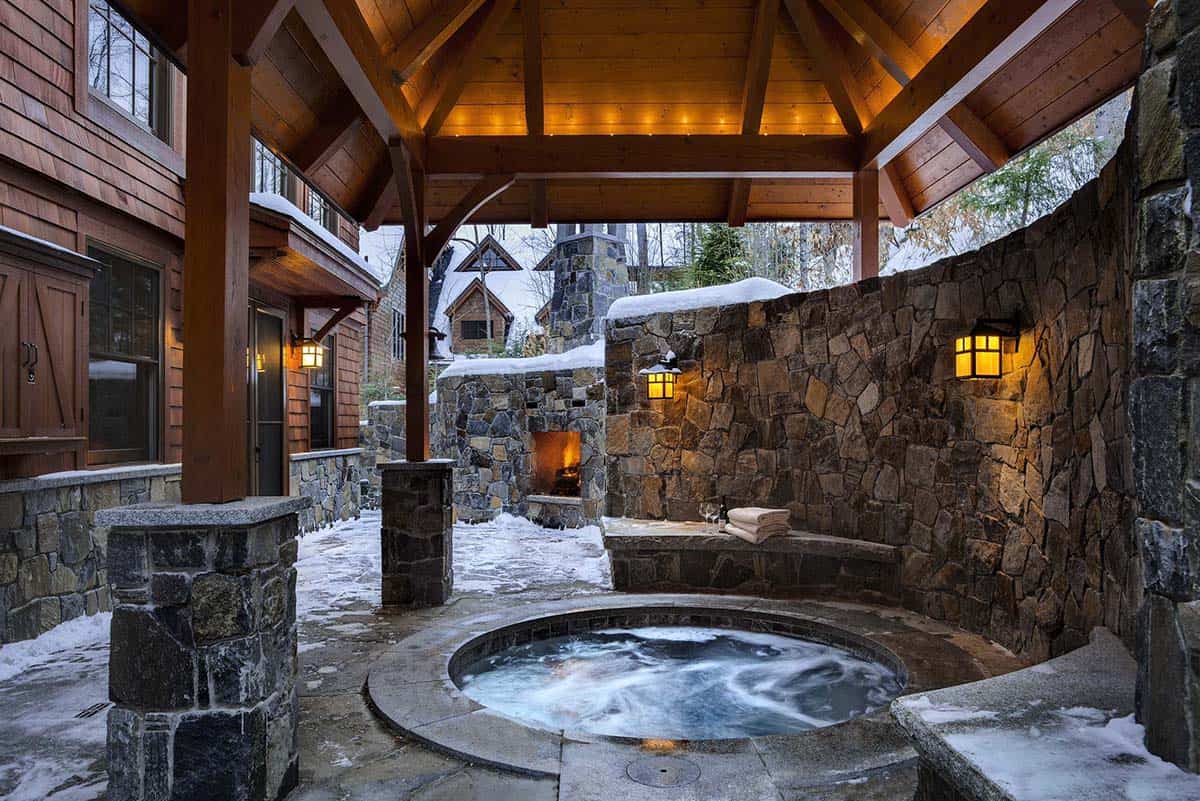 Sunken stone hot tub under a timber pavilion with a curved fieldstone wall, lantern sconces and an outdoor fireplace