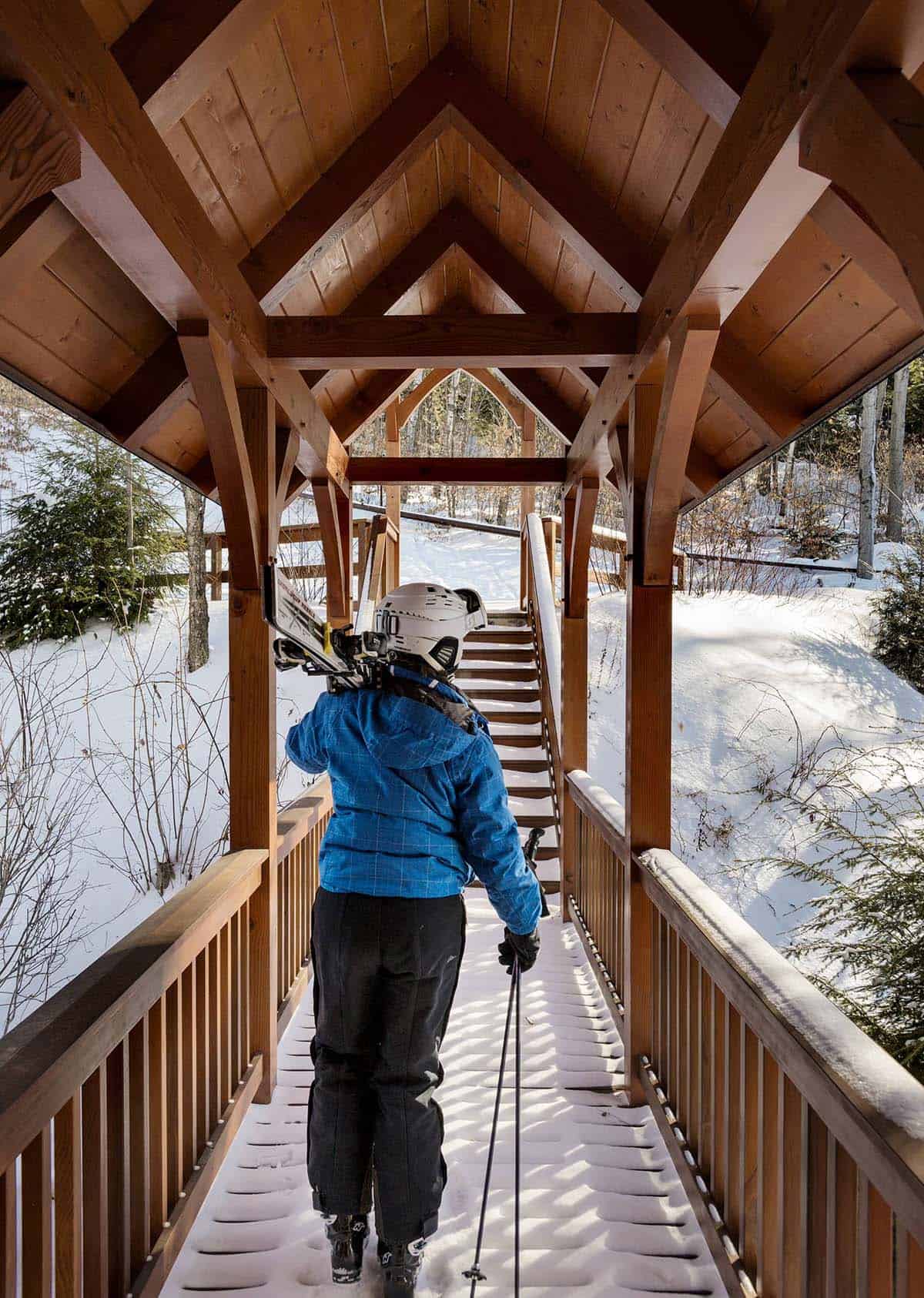 Skier carrying skis across a covered timber bridge toward stairs in the snowy woods