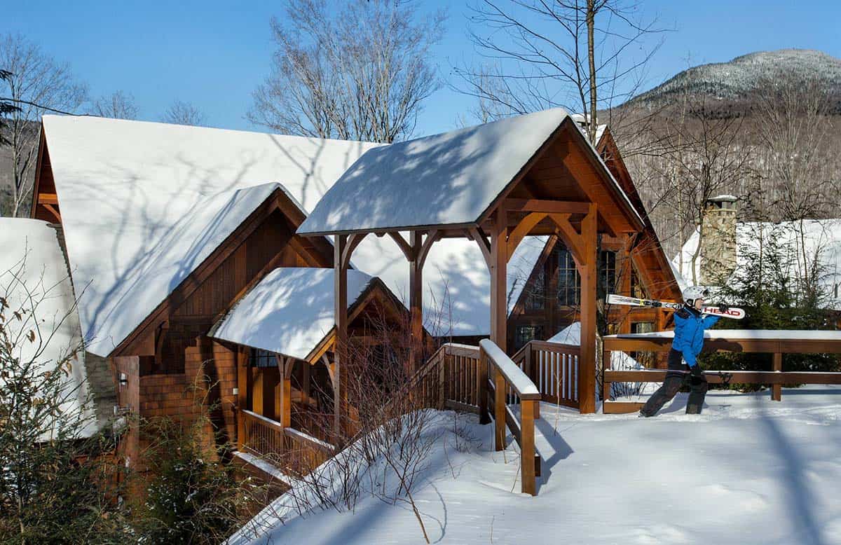Covered timber-frame bridge connecting the snow-covered home to a ski trail, with a skier and mountain beyond