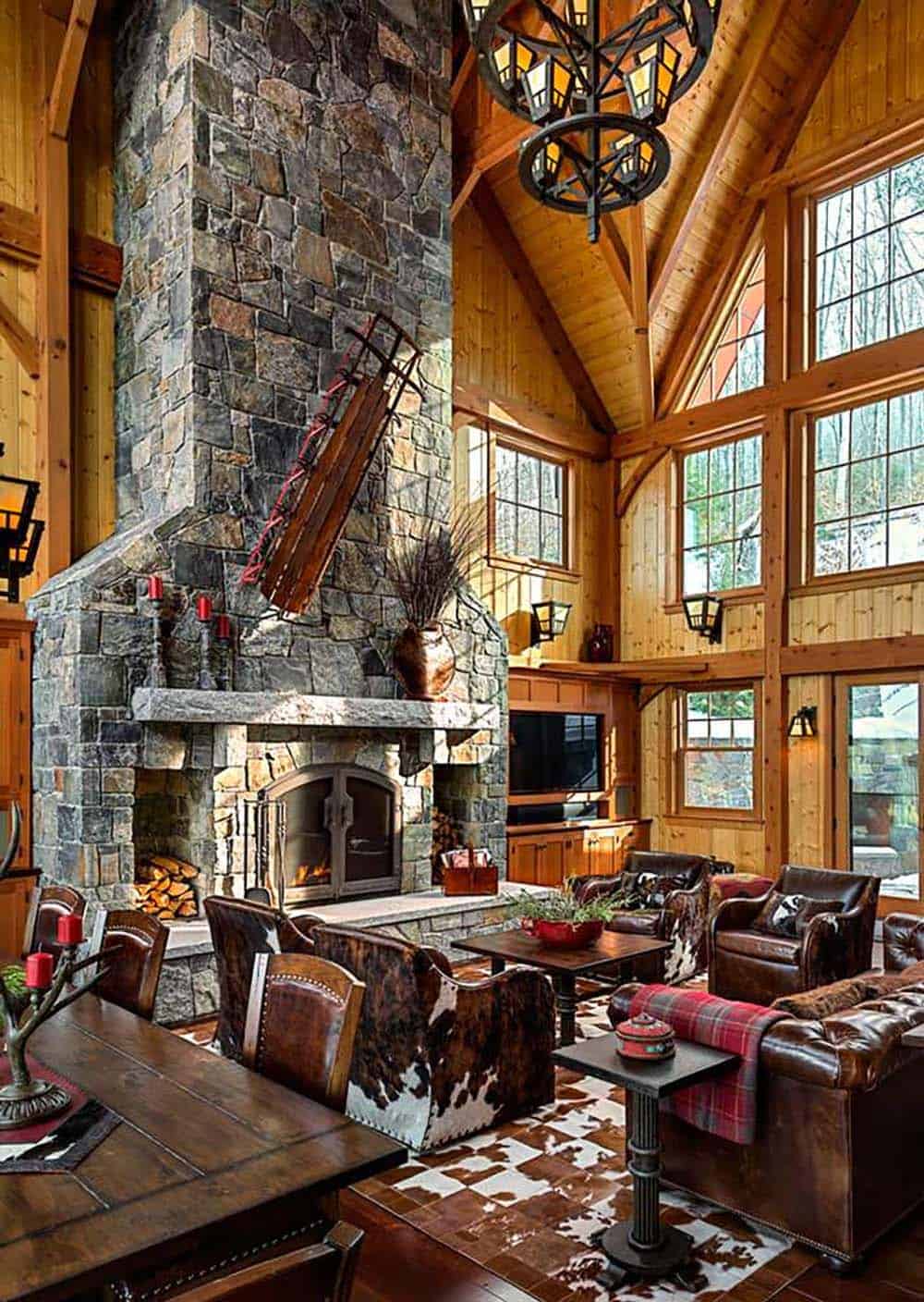 Great room with a towering stone fireplace, cowhide and leather chairs and tall timber-framed windows