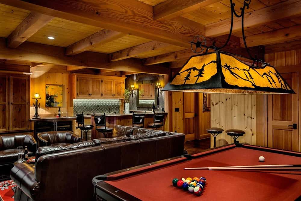 Lower-level lounge with a leather sectional facing a cowhide-stool bar, beside a pool table lit by a pine-tree silhouette pendant