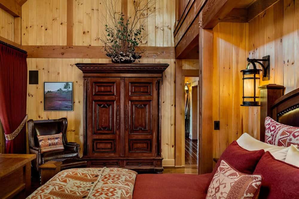 Bedroom with knotty pine walls, a carved wood armoire, leather wingback chair and lantern sconce beside a red upholstered bed