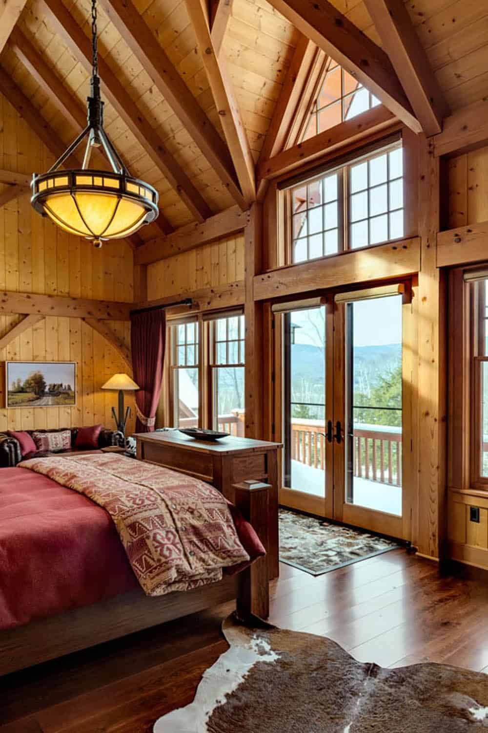 Primary bedroom with a vaulted timber-frame ceiling, red bedding, cowhide rug and glass doors leading to a snowy deck with mountain views